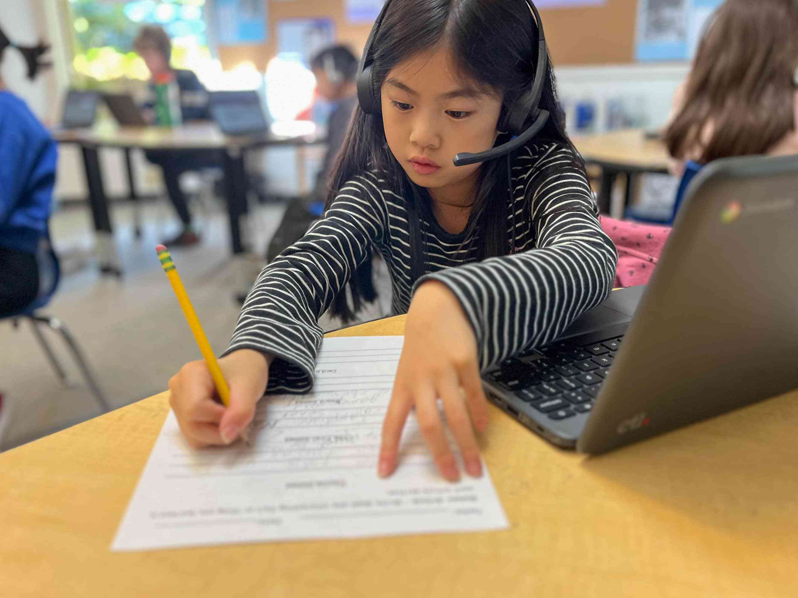 A child writing on a worksheet with a headset on.